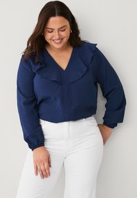 Blusa blu navy con scollo a V e dettagli a rouches lungo le spalle. Maniche lunghe con polsini elasticizzati, tessuto texturizzato, abbinata a pantaloni bianchi.