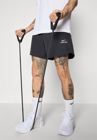 Svarta Nike Pro-shorts, grå och vita sneakers, vita ankelsockor, hållandes svarta motståndsband. Noterbara tatueringar synliga på benen.