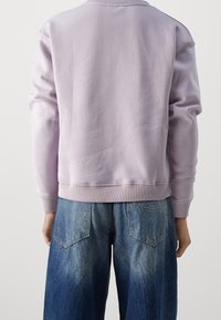 Lilac sweatshirt dengan hem dan manset berenda, dipadukan dengan celana jins denim biru longgar yang memiliki aksen pudar dan patch bermerek di belakang.