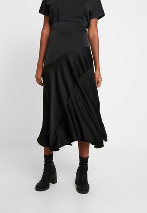 Frau trägt einen schwarzen asymmetrischen Midirock aus einer Mischung aus matten und satinierten Stoffen, kombiniert mit schwarzen Stiefeletten mit Absatz.