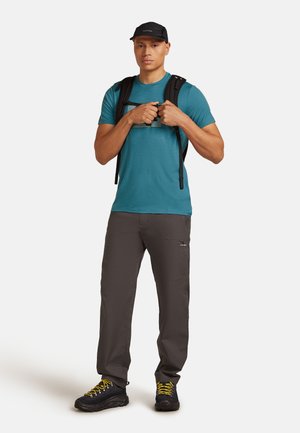 Teal T-shirt met korte mouwen, grijze cargo broek, zwarte wandelschoenen met gele veters, zwarte pet en een zwarte rugzak.