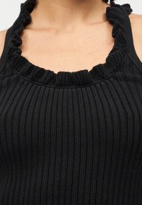 Κοντινό πλάνο στο άνω μέρος του στήθους ενός ατόμου που φοράει μαύρο ριμπ πλεκτό τοπ με βολάν κατά μήκος του λαιμοκοψίματος και των τιράντων.