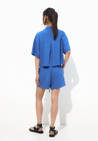 Camisa de lino azul cortada con mangas cortas, que presenta un pliegue en la parte posterior. Combinada con pantalones cortos azules y sandalias negras de tiras. Fondo liso.