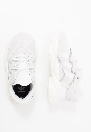 Paire de baskets blanches Adidas avec semelles épaisses, panneaux en suède et en mesh, et logo Adidas visible à l'intérieur de la chaussure gauche.
