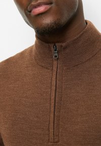 Brauner Pullover mit Reißverschluss, hohem Kragen und strukturiertem Strickstoff, mit einem schwarzen Reißverschluss vorne.