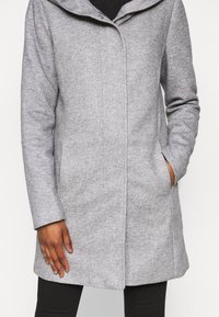 Abrigo texturizado de longitud media en gris claro con botones ocultos, llevado sobre pantalones negros, mano en el bolsillo derecho, sobre un fondo neutro.