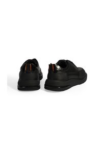 Des chaussures de sport noires avec une tige en cuir lisse et texturé. Elles possèdent une semelle en caoutchouc et un accent orange à l'arrière. Vue arrière montrée.