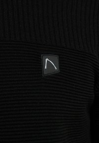 Schwarzer gestrickter Pullover mit Rippstruktur, ausgestattet mit einem kleinen schwarzen Patch, der eine weiße Lin Zeichnung zeigt.