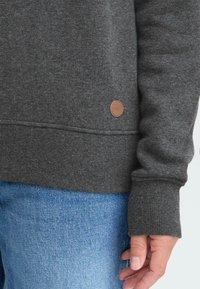 Kullgrå genser med ribbet tekstur, rund halslinning og lærlogo-patch nederst. Blå denimjeans delvis synlige.