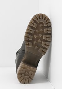 Semelle de botte noire avec un motif de semelle profond, comprenant des croix et des formes géométriques, inclinée contre un fond blanc en coin.