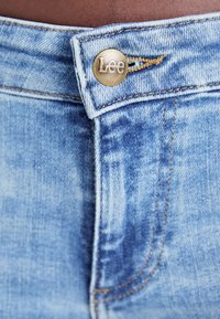 Jean en denim bleu clair avec un bouton en métal portant le logo "Lee", des coutures contrastantes orange, et un design classique à cinq poches.
