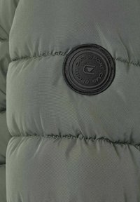 Quiltat mörkgrönt tyg med en svart rund emblem märkt "CMS SPORT" sydd på vadderat material.