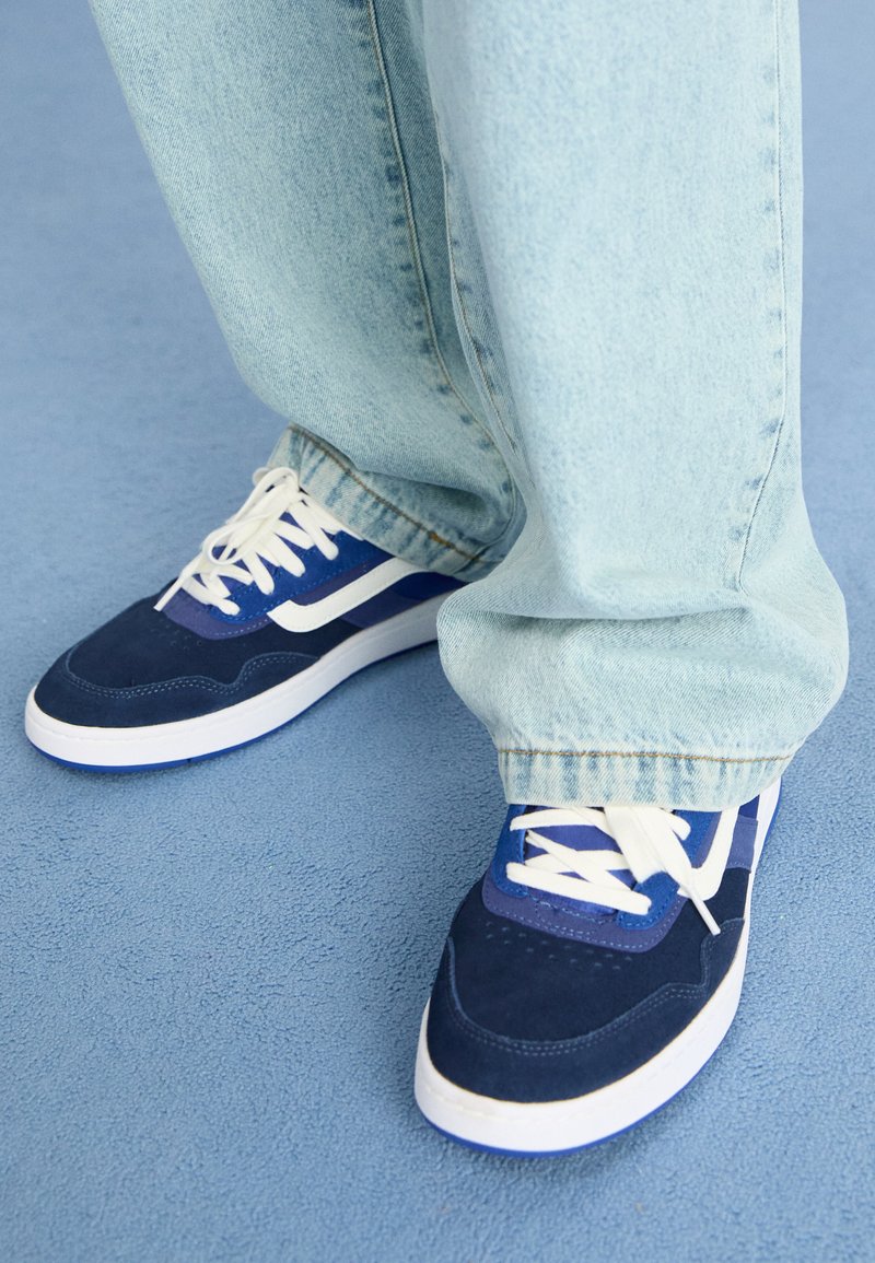 Baskets en suede bleu avec des lacets blancs, arborant un motif de vagues bleu et blanc sur le côté, associées à un jean en denim bleu clair.