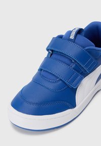 Basket synthétique bleue avec des sangles Velcro, accents blancs, texture lisse, bout arrondi et intérieur rembourré. Présente une étiquette de logo sur le talon.