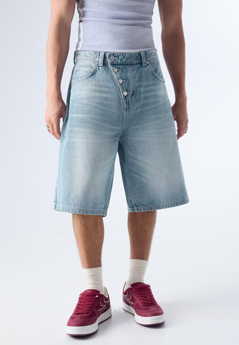 Personne portant un short en denim bleu clair jusqu'aux genoux, un débardeur blanc côtelé, des chaussettes blanches et des baskets marron, debout sur un fond uni.