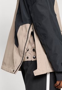 Bikola jakna v bež in temno sivi barvi, ki ima zadrgo, gumbke na pritisk in teksturirano tkanino s kontrastnimi paneli.