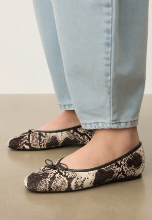 Kājās valkātas baleta kurpes ar čūskas ādas rakstu, saskaņotas ar gaiši zilām džinsu biksēm, stāvot uz gludas grīdas.