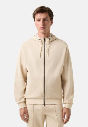 Beige zip-up hoodie med justerbar hætte, ribbede ærmer og kant, med en glat tekstur og minimalistisk design.