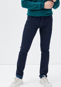 Jeans slim-fit blu navy con una sottile texture, dotati di tasche frontali, chiusura con zip e orli arrotolati sopra scarpe da ginnastica bianche.