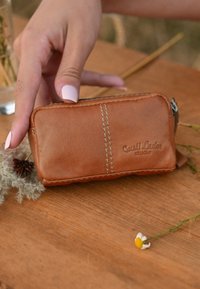 Smeđi kožni zippered novčanik s glatkom teksturom, s vidljivim šavovima i logotipom urezanim sa strane. Mjeri približno 10 cm x 5 cm.