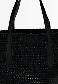 Sac fourre-tout tissé noir avec des poignées en cuir lisse. Texture distinctive grâce aux matériaux entrelacés. Forme élégante et structurée. Aucune quincaillerie visible.