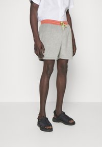Pantalones cortos de color gris claro con una cinturilla de cuerda naranja, hechos de un material suave. Combinados con sandalias negras con los dedos al aire y correas.