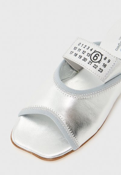 Ασημένιο σανδάλι με ανοιχτά δάχτυλα, γκρι ανοιχτές λεπτομέρειες και λευκό λουράκι που φέρει μαύρους αριθμούς που περιστρέφονται γύρω από το μέγεθος 6 σε έντονο κυκλικό σχέδιο.