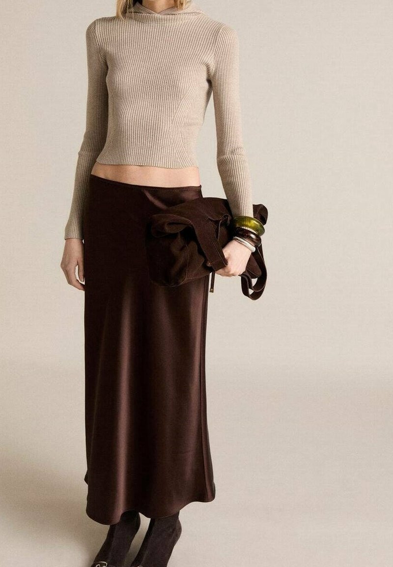 Femme portant un crop top à manches longues côtelé beige, une jupe longue en satin marron, tenant un sac en daim marron, et portant des bottines noires et des bracelets.