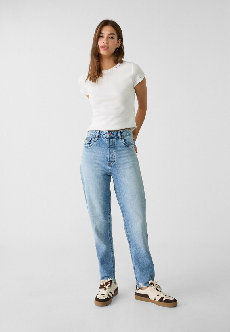 T-shirt blanc à manches courtes avec des accents de broderie, associé à un jean taille haute bleu clair et des baskets crème avec des motifs bruns.