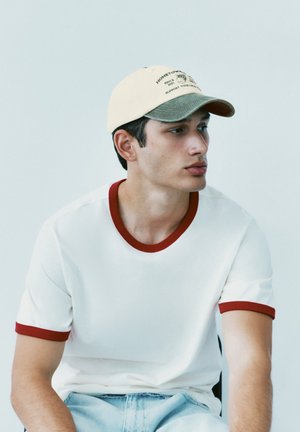 Jeune homme portant un t-shirt blanc avec une bordure rouge et une casquette beige et verte, regardant sur le côté sur un fond clair uni.
