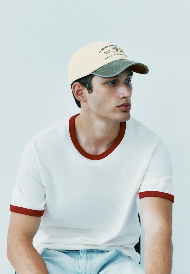 Jeune homme portant un t-shirt blanc avec une bordure rouge et une casquette beige et verte, regardant sur le côté sur un fond clair uni.