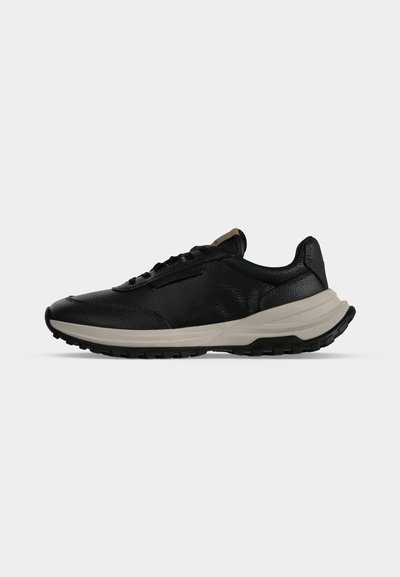 Μαύρο sneaker χαμηλού πάνω μέρους με υφή δέρματος, λευκή χοντρή σόλα και μαύρη εξωτερική σόλα με πέλμα, εμφανιζόμενο από την εξωτερική πλευρά σε γκρι φόντο.