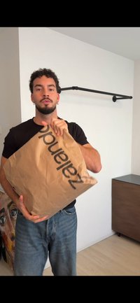 Καφέ χάρτινη τσάντα με μαύρο τυπωμένο λογότυπο που κρατιέται από ένα άτομο, με ζαρωμένη υφή. Στο παρασκήνιο διακρίνεται μια ξύλινη ντουλάπα.