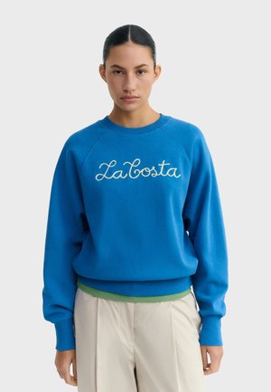 Žena v modré mikině s nápisem "La Costa", béžovými kalhotami a zeleným páskem stojí před jednoduchým světlým pozadím.