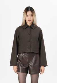 Ung kvinna med långt blont hår bär en mörkbrun knappad kort jacka, bruna lädershorts och genomskinliga svarta strumpbyxor, stående mot vit bakgrund.