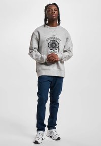 Felpa grigia con un design del logo nero sulla parte anteriore, abbinata a jeans blu scuro e sneakers bianche. Tessuto morbido e vestibilità comoda.