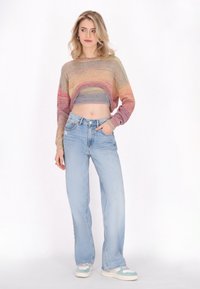 Sokszínű kötött, crop pulóver ribbelt végű, világoskék, bő szárú farmernadrággal és két színű sportcipőkkel párosítva. A modell az egyik kezével a zsebében áll.