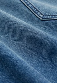 Tela de mezclilla en azul desgastado, con una textura sutil que presenta patrones de tejido diagonales y un detalle de bolsillo visible en la esquina superior izquierda.