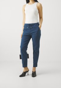 Marineblaue, schmal zulaufende Hose mit elastischem Bund, zwei Seitentaschen und lässiger Passform, kombiniert mit schwarzen, offenen High Heels und einer kleinen Tasche.