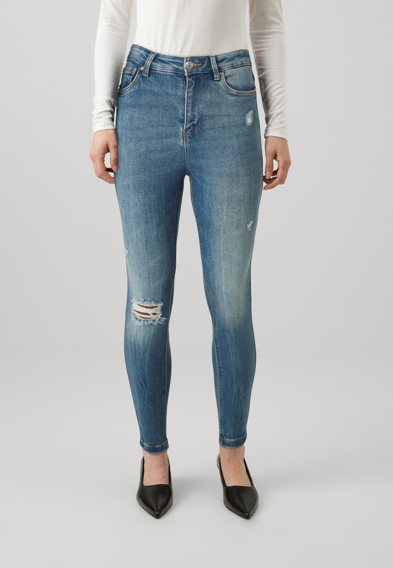 Jeans de denim azul de tiro alto con detalles de rodilla rasgada, con un corte ajustado y un ligero desgastado. Combinados con tacones negros de punta afilada.