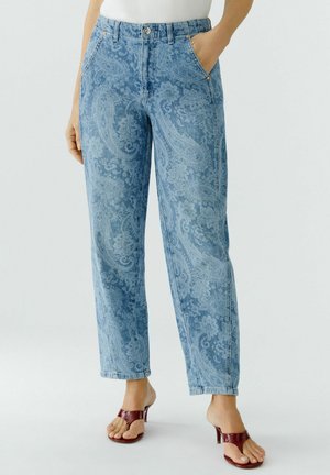 Frau trägt hoch taillierte blaue Jeans mit Paisley-Muster, weißes Oberteil und bordeauxrote Sandalen mit Absatz, steht mit Händen in den Taschen.