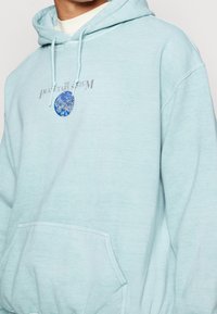 Ljusblå bomullshoodie med en framficka, som har grafisk texten "DE PLANETÄRA SYSTEMET" och en blå cirkulär design.