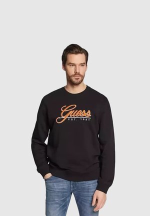 Mann trägt schwarzen Pullover mit der Aufschrift "Guess EST. 1981", kombiniert mit blauer Jeans, steht vor einem schlichten hellgrauen Hintergrund.