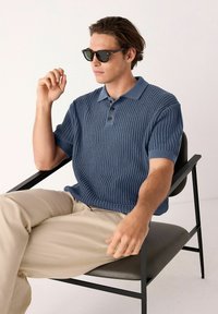 Polo shirt tricotat texturat bleu cu mâneci scurte, cu trei butoane maro la guler, purtat cu pantaloni bej, așezat pe un scaun negru.