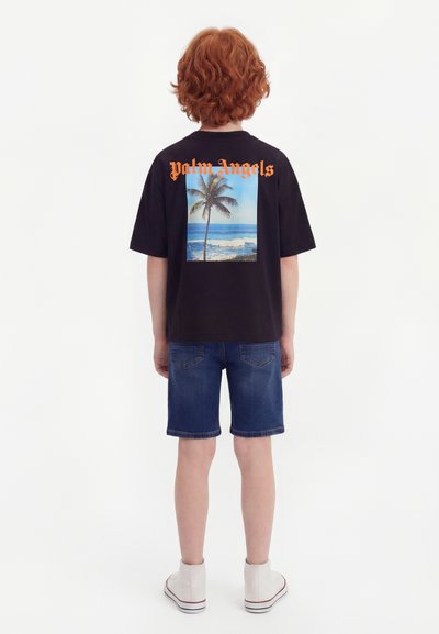 Παιδί με κόκκινα σγουρά μαλλιά στέκεται γυρισμένο από πίσω, φορώντας μαύρο μπλουζάκι με εκτύπωση παραλίας με φοίνικα και πορτοκαλί κείμενο, τζιν σορτς και λευκά αθλητικά παπούτσια.