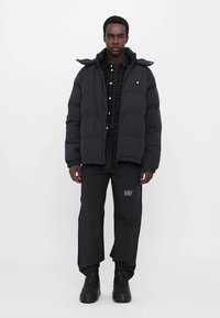 Black puffer jacket with a hood, featuring a quilted texture. Paired with black pants and a checkered shirt, worn with black boots.