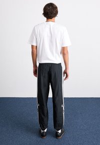 Černé sportovní kalhoty s bílými bočními pruhy, elastickým pasem a decentním logem na dolní části nohy, v kombinaci s jednoduchým bílým tričkem s krátkým rukávem.