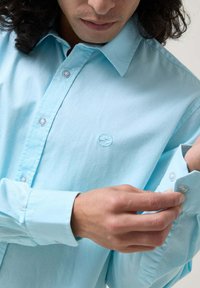 Camisa azul clara de botones con un cuello estructurado, mangas largas y un logo bordado. La tela tiene un acabado suave y ligeramente texturizado.