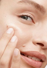 Gros plan sur un visage à la peau lisse et claire ; une main touche la joue. Le visage présente des lèvres de couleur claire, un grain de beauté et une forme de sourcils naturelle.