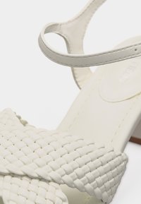 Sandale blanche avec bride avant tressée et fine bride à la cheville sur un petit talon bloc, présentée en détail rapproché.